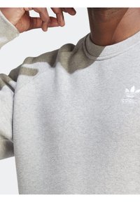 Šedá mikina s kulatým výstřihem, vyrobená z texturované látky a s jemnými tónovými akcenty. Malé bílé logo na levé části hrudi.