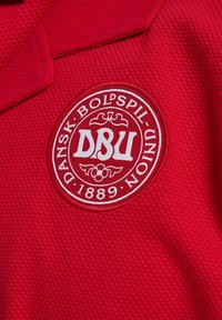 Czerwona teksturowana tkanina z białym okrągłym emblematem z napisem "DANSK BOLDSPIL UNION 1889" oraz dużymi inicjałami "DBU" na środku.