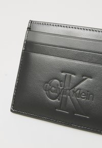 Juodas odinis kortelių laikiklis su įspaustu Calvin Klein logotipu, penkiomis kortelių lizdais ir siūlėmis aplink kraštus. Textūruotas paviršius.