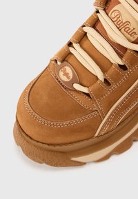 Hnedé semišové tenisky s béžovými akcentmi, hrubou gumovou podrážkou a svetlými šnúrkami. Obsahujú prešité detaily a dizajnovú nášivku s logom na boku.