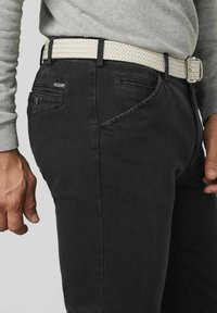 Zwarte denim broek met een slim fit, voorzien van een gevlochten witte riem en contrasterende stiksels. De zijaanzicht benadrukt de zak- en riemdetails.