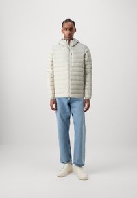 Jachetă puffer de culoare bej deschis, cu cusături orizontale, fermoar frontal și glugă. Asortată cu blugi albaștri deschis și adidași albi. Design simplu.