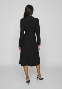 Long black knit dress with a fitted bodice and flared pleated skirt. Features a matching belt and long sleeves, made from soft material.
