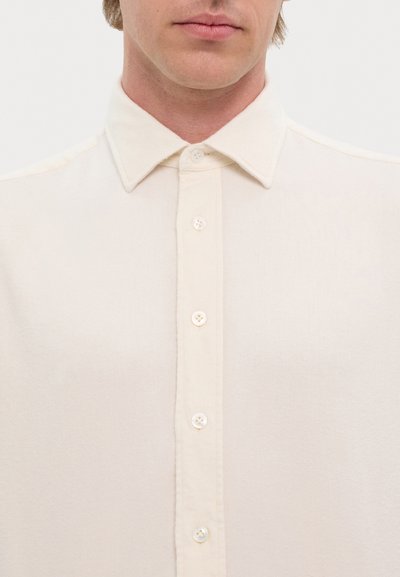 Camisa de botón color beige claro, hecha de un tejido texturizado, con un cuello clásico y siete botones en la parte delantera.