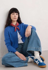 Blå stickad cardigan med knappar, bärs över en vit topp. Utrustade jeansbyxor och blå sneakers med vita detaljer. Röd scarf knuten runt halsen.