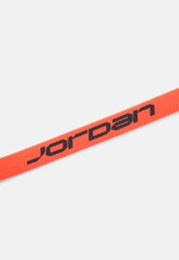 Raudonas pintas diržas su juodu "Jordan" logotipu. Plokščias dizainas, tekstūruotas paviršius, tinkantis naudoti kaip diržas ar aksesuaras.