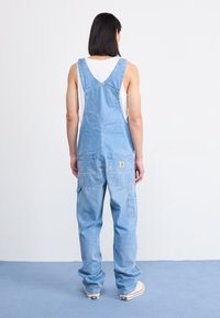 Blaue Denim-Latzhose mit lockerer Passform, ausgestattet mit großen Taschen und verstellbaren Trägern, getragen über einem weißen Tanktop.