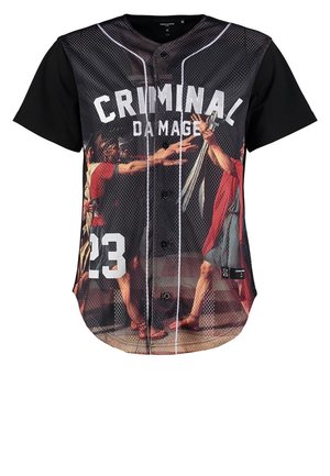 Homme portant un t-shirt graphique noir et gris avec l'inscription « CRIMINAL DAMAGE 23 », un jean gris, des bottes noires, tenant un grand sac de sport noir.
