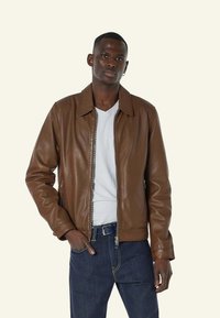 Bruine leren jacket met een rits aan de voorkant, puntige kraag en zijzakken, gedragen over een wit T-shirt en blauwe jeans. Glad textuur.