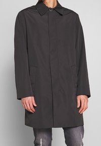 Mand i en sort langærmet knapjakke med sidelommer over slidte grå jeans foran en ensfarvet baggrund.