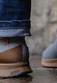 Zapatilla gris y marrón con parte superior texturizada, que incluye una etiqueta resistente al agua. Suela robusta en un color marrón claro contrastante.