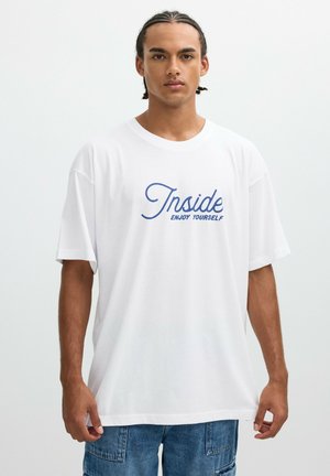 Joven con cabello trenzado que lleva una camiseta blanca con texto azul que dice "Inside Enjoy Yourself" y pantalones cortos de mezclilla azules, de pie contra un fondo simple.