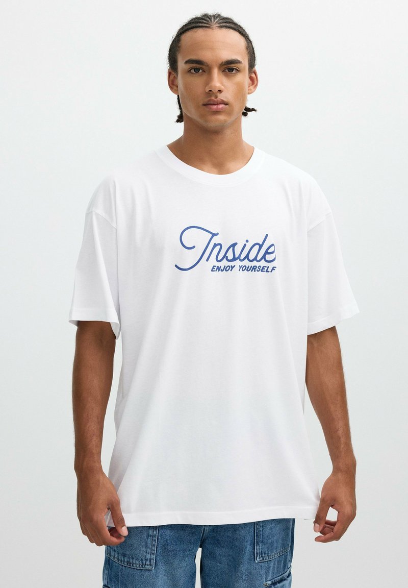 Joven con cabello trenzado que lleva una camiseta blanca con texto azul que dice "Inside Enjoy Yourself" y pantalones cortos de mezclilla azules, de pie contra un fondo simple.