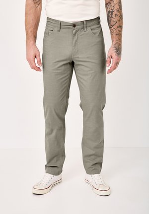 Mann trägt hellgraue Freizeithose und weiße Turnschuhe, steht vor neutralem Hintergrund, sichtbar von der Taille bis zu den Füßen.