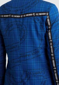 Veste à carreaux bleue avec des motifs textuels noirs et un ruban latéral contrastant portant l'inscription « RESSENS TON ÉNERGIE ». Le matériau semble texturé avec un design structuré.