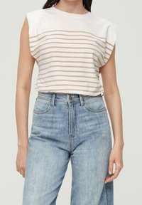Žena v bílém bezrukávovém topu s béžovými vodorovnými pruhy a světle modrých džínách s vysokým pasem na jednobarevném pozadí.