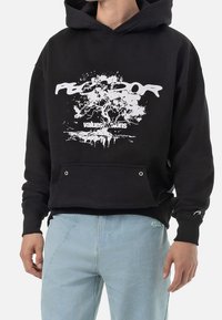 Svart hoodie med en framgrafik av ett träd och vit text. Har en känguruficka och ribbstickade muddar. Stylad med ljusblå byxor.