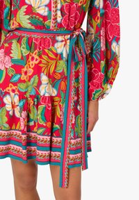 Rotes Blumenkleid mit lebendigen Mustern, langen Ärmeln, einem Bindegürtel und einem Rüschensaum, mit türkisfarbenen Akzenten und Details.