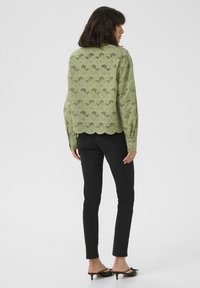 Chemise vert clair à bords festonnés avec un motif floral découpé au laser, associée à un jean skinny noir et des mules à talons noires.