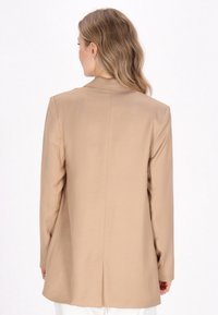 Beige bleiseri, jossa on rakenteellinen muotoilu, pitkät hihat ja yksittäinen halkio takana. Tasainen kangas, jossa on minimaalinen näkyvä detaljointi.