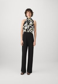 Svart jumpsuit med halterneck, med et blomstermønstrete liv i svart, hvitt og gult. Stoffet har en glatt tekstur.
