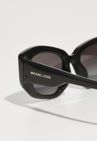 Lieli melni saulesbrilles ar ģeometrisku rāmi, gradiento lēcu un zelta "MICHAEL KORS" logotipu uz sāniem.