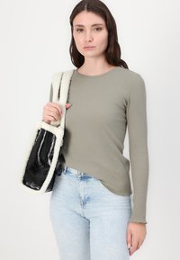 Duga majica s rebrastim uzorkom u maslinasto zelenoj boji, u kombinaciji s svijetloplavim trapericama. U jednoj ruci drži torbu s umjetnim krznom i sjajnim crnim panelom.