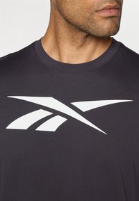 Melnas sporta t krekls ar lielu baltu logo dizainu uz krūtīm. Audums ir gluds, un apkakle ir apaļa.