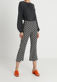 Top nero a maniche lunghe con pieghe, pantaloni cropped a righe nere e bianche, e mule arancioni con tacco blocco. Contrasto testurale nei materiali.
