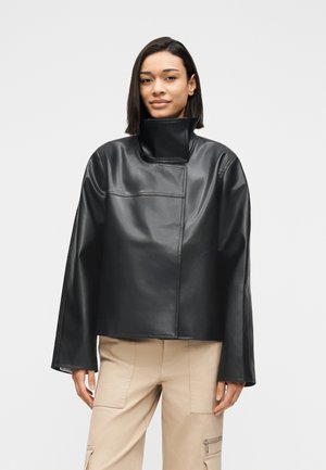 Femme portant une veste en cuir noire à col haut avec des manches larges et un pantalon cargo beige sur un fond blanc uni.