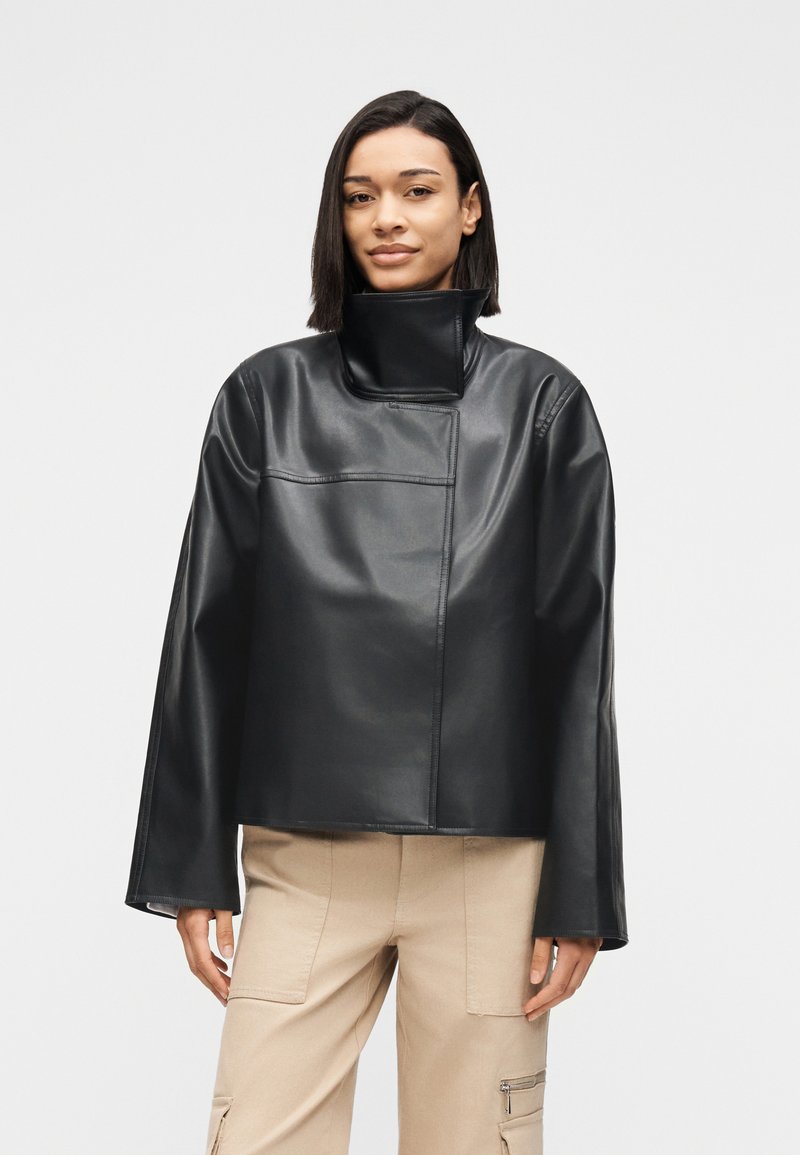 Γυναίκα που φοράει μαύρο δερμάτινο μπουφάν με ψηλό γιακά και φαρδιά μανίκια, μαζί με μπεζ παντελόνια καμπάνιας, πάνω σε απλό λευκό φόντο.