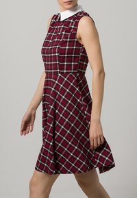Červené kostkované šaty s bílým límečkem, přiléhavým trupem, rozšířenou sukní, bez rukávů a texturovanou látkou s bílými a černými pruhy.