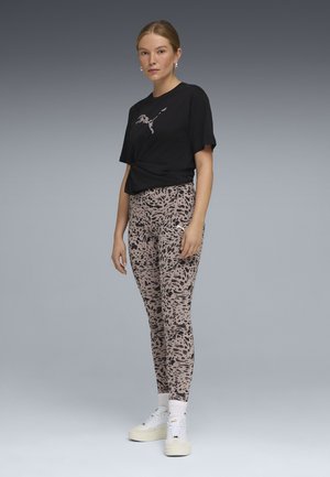 Mujer de pie con camiseta negra de Puma, leggings beige con manchas negras, calcetines blancos y zapatillas blancas Puma sobre fondo gris.