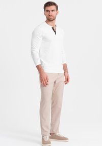 Vit långärmad henley-tröja med svart knappslå, ihop med ljusbeiga byxor och beigefärgade sneakers. Enkel, modern passform.