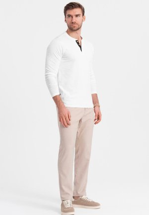 Camisa henley de manga larga blanca con placket de botones negros, combinada con pantalones beige claros y zapatillas beige. Fit simple y moderno.