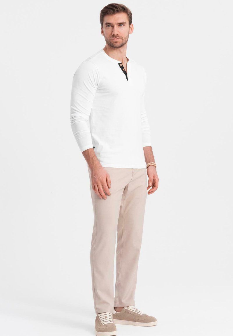 Vit långärmad henley-tröja med svart knappslå, ihop med ljusbeiga byxor och beigefärgade sneakers. Enkel, modern passform.