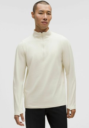 Helles cremefarbenes Langarm-Top mit Viertel-Reißverschluss aus weichem Stoff, mit hohem Kragen und tailliertem Schnitt, glatter Textur und minimalen Nähten.