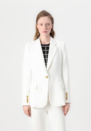Blazer blanco hecho de tela de sastrería, con un solo botón dorado, dos bolsillos frontales y acentos de botones dorados en las mangas.