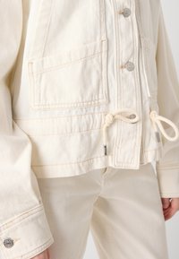Beige denimjakke med to frontlommer, sølvknapper og snor i nederkanten. Myk tekstur, synlige sømdetaljer, avslappet passform.