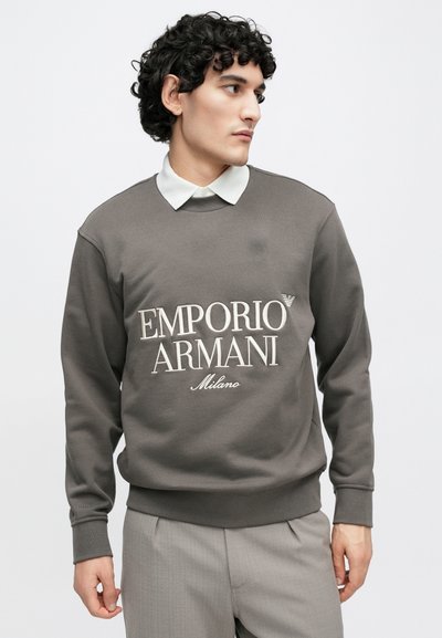 Hnedá mikina s nápisom "Emporio Armani Milano" potlačeným na bielo. Má okrúhly výstrih a žebrované manžety, nosená cez bielu košeľu s golierom.