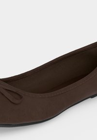 Ballerine marron avec un petit nœud sur le dessus et un bout arrondi, dotée d'une semelle noire basse.