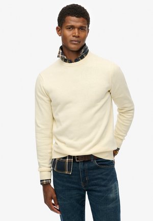 Suéter de punto color crema con cuello redondo, superpuesto a una camisa de cuadros, combinado con jeans azul oscuro y un cinturón marrón.