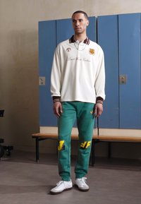 White long-sleeve shirt with brown collar and logo, paired with green pants featuring yellow accents and text, worn with white sneakers.