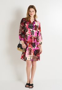 Vestido floral colorido con mangas largas, escote en V y cintura fruncida. La tela presenta patrones abstractos en rosa, marrón y negro. Combinado con un bolso pequeño.