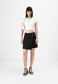 Donna con capelli lisci e scuri, che indossa un top corto bianco, una gonna avvolgente nera con dettagli dorati, sandali neri con tacco alto e porta una borsa a spalla rosa chiaro.