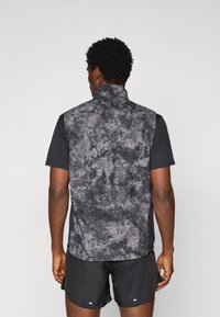 Mann trägt eine ärmellose Weste mit schwarz-grauem Muster über einem schwarzen Kurzarmhemd und schwarzen Sport Shorts, steht vor einem schlichten Hintergrund.