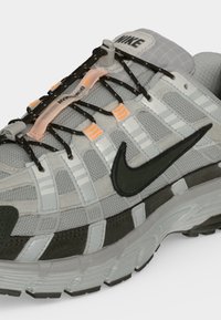 Γκρι και μαύρο αθλητικό παπούτσι Nike με ανακλαστικές λευκές ρίγες, μαύρα κορδόνια με πορτοκαλί λεπτομέρειες και ορατό λογότυπο Nike στο πλάι και στη γλώσσα.