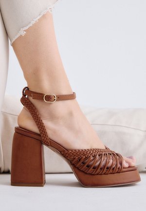 Pie con sandalia de plataforma de cuero marrón con tira trenzada y hebilla en el tobillo, sobre pantalones beige y fondo claro.