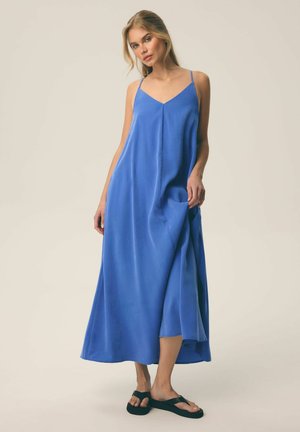 Kobieta ubrana w bezrękawową niebieską sukienkę maxi z cienkimi ramiączkami i czarne sandały, stojąca na tle jednolitego jasnego tła.