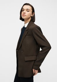 Νεαρό άτομο με κοντά σκούρα μαλλιά, φορώντας καφέ σακάκι πάνω από λευκό πουκάμισο με γιακά και σκούρο πουλόβερ, ποζάρει μπροστά σε απλό φωτεινό φόντο.